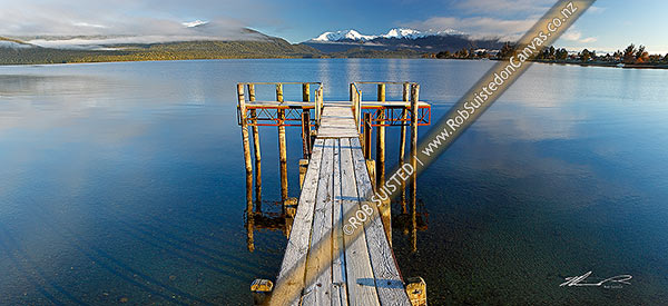Photo of Lake Te Anau and lakeside jetty, with winter snow capped Murchison Mountains and Fiordland National Park beyond. Calm early morning panorama, Te Anau, Southland, Southland Region, New Zealand (NZ)