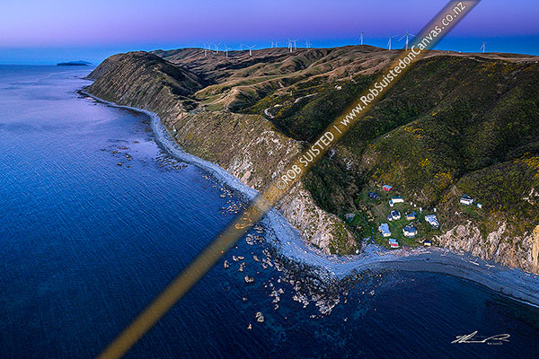 Photo of Mill Creek Wind Farm turbines above Smiths Bay and baches on the Makara Beach coastline. Looking north towards Mana and Kapiti islands in the sunset after glow. Aerial view, Makara Beach, Wellington City, Wellington Region, New Zealand (NZ)